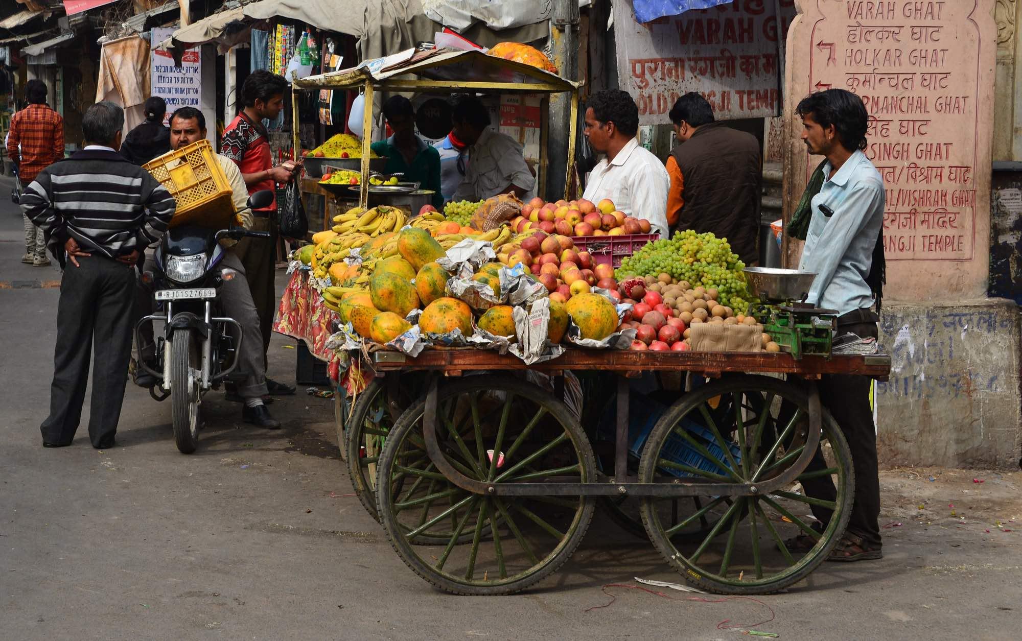 A street market in Pushkar, Rajasthan — a fruit vendor's handcart on the roadside with shoppers and a parked motorcycle alongside.